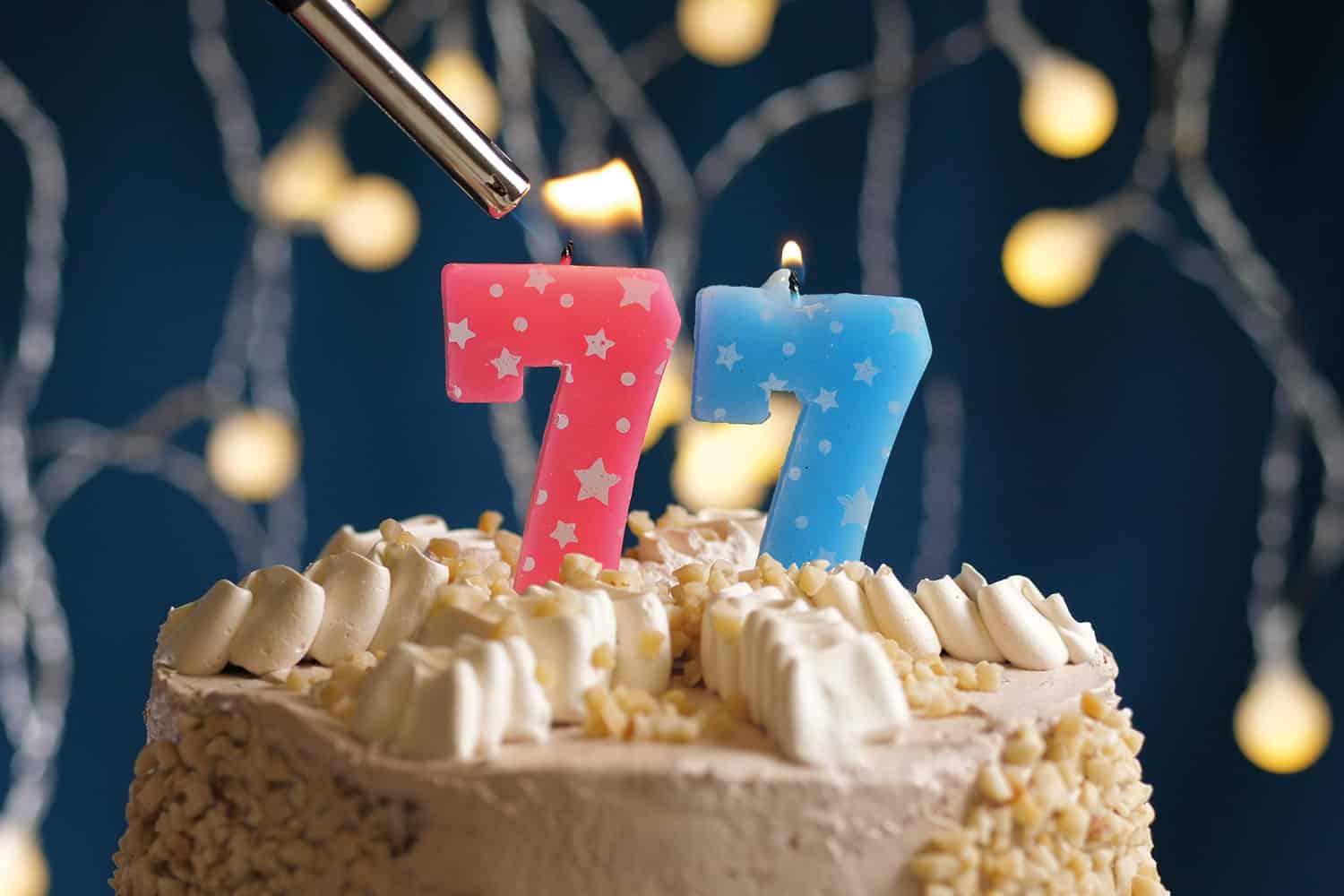 creamy birthday cake with two numeric candles making the number ‘77’ being lit against a backdrop of party lights