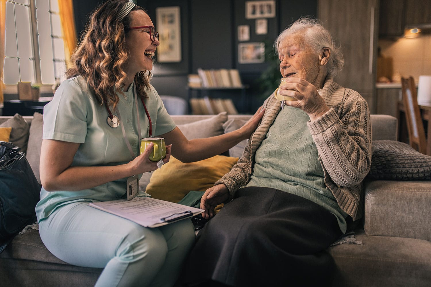 photo of young woman carer having a cup of tea and a laugh with an older woman on her sofa