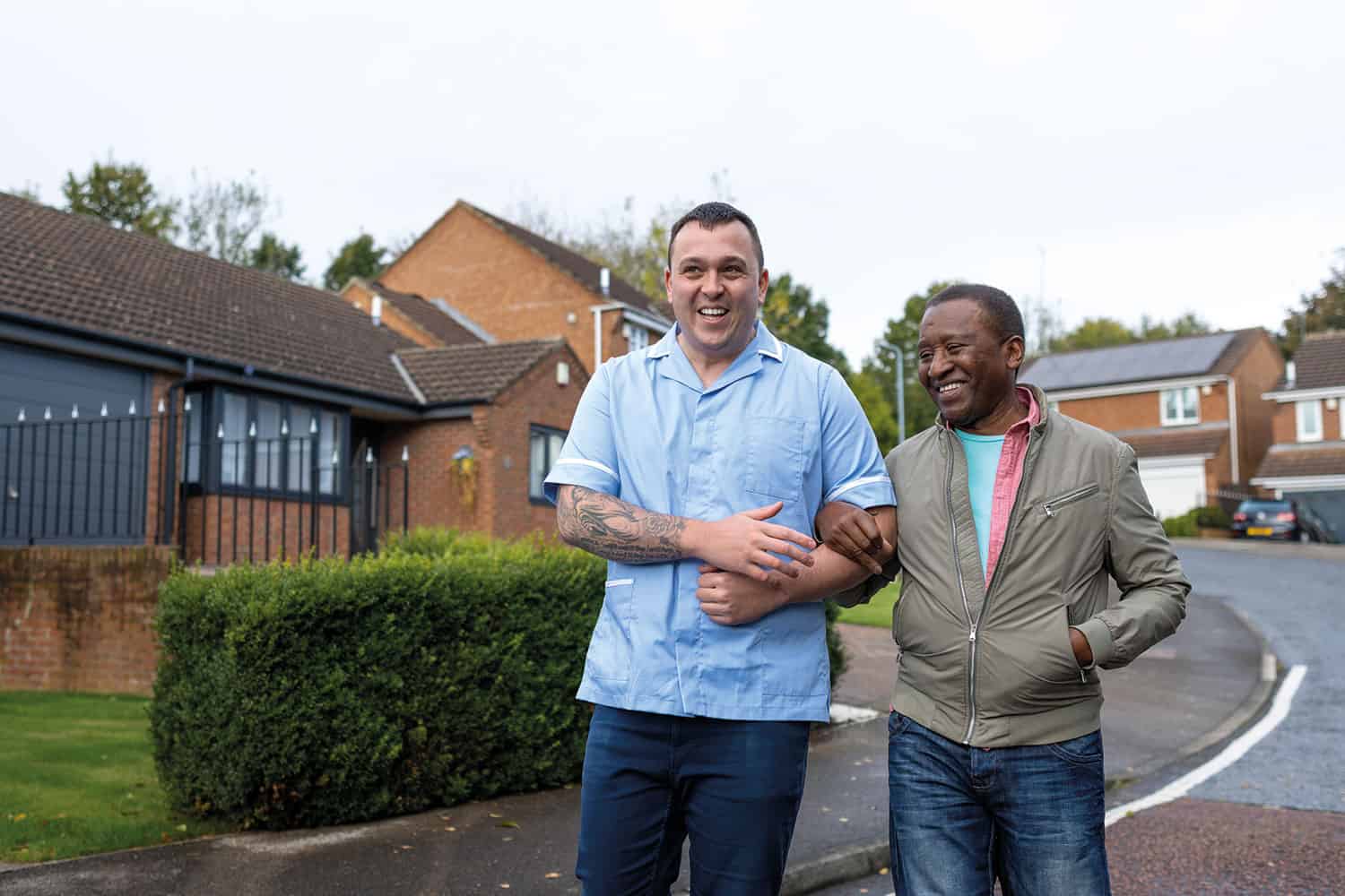 photo of male carer in blue medical tunic arm-in-arm with older man, both smiling, walking outside past housing