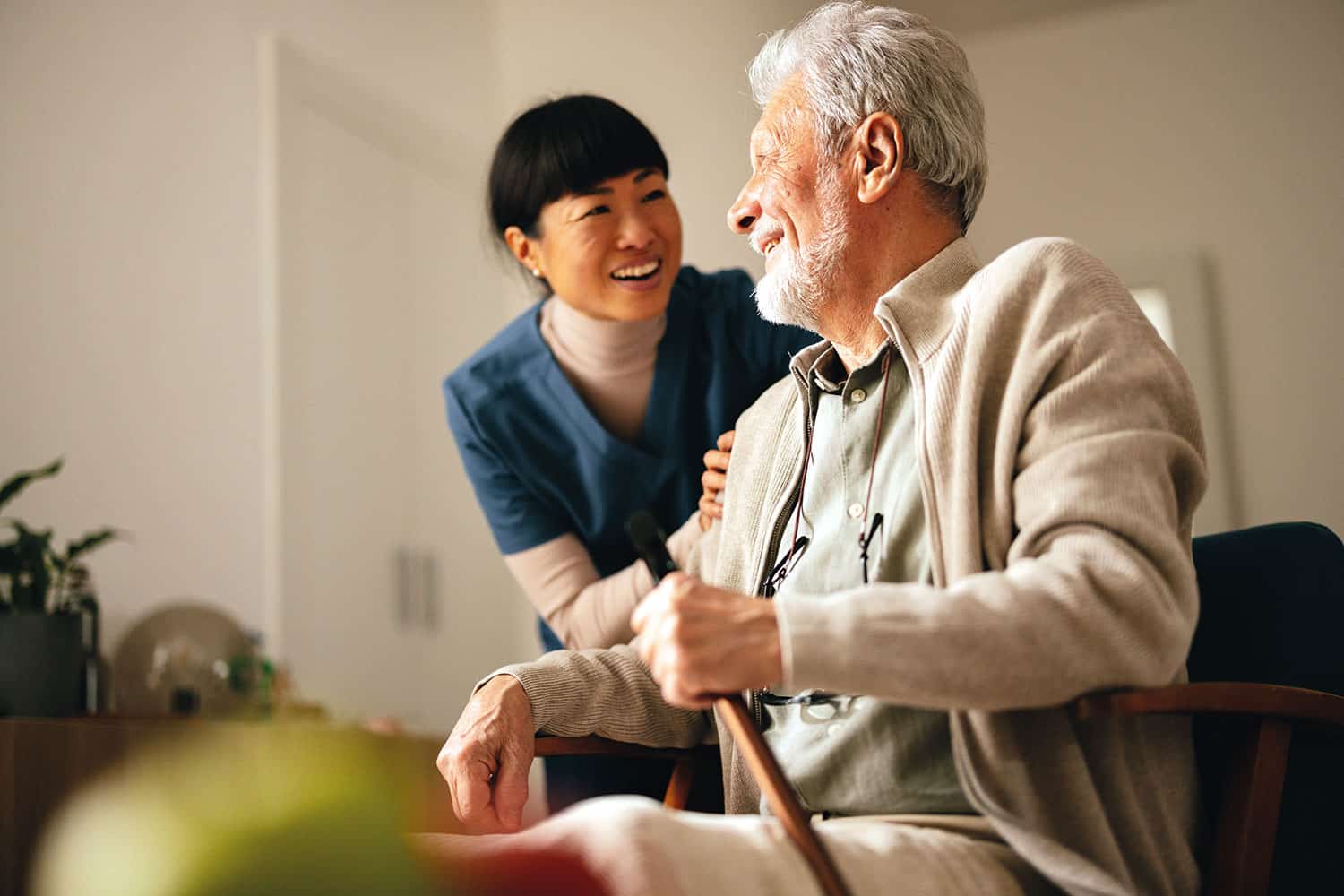 side-on shot of older man seated holding walking stick looking up at female carer, who is leaning in over his shoulder, both smiling