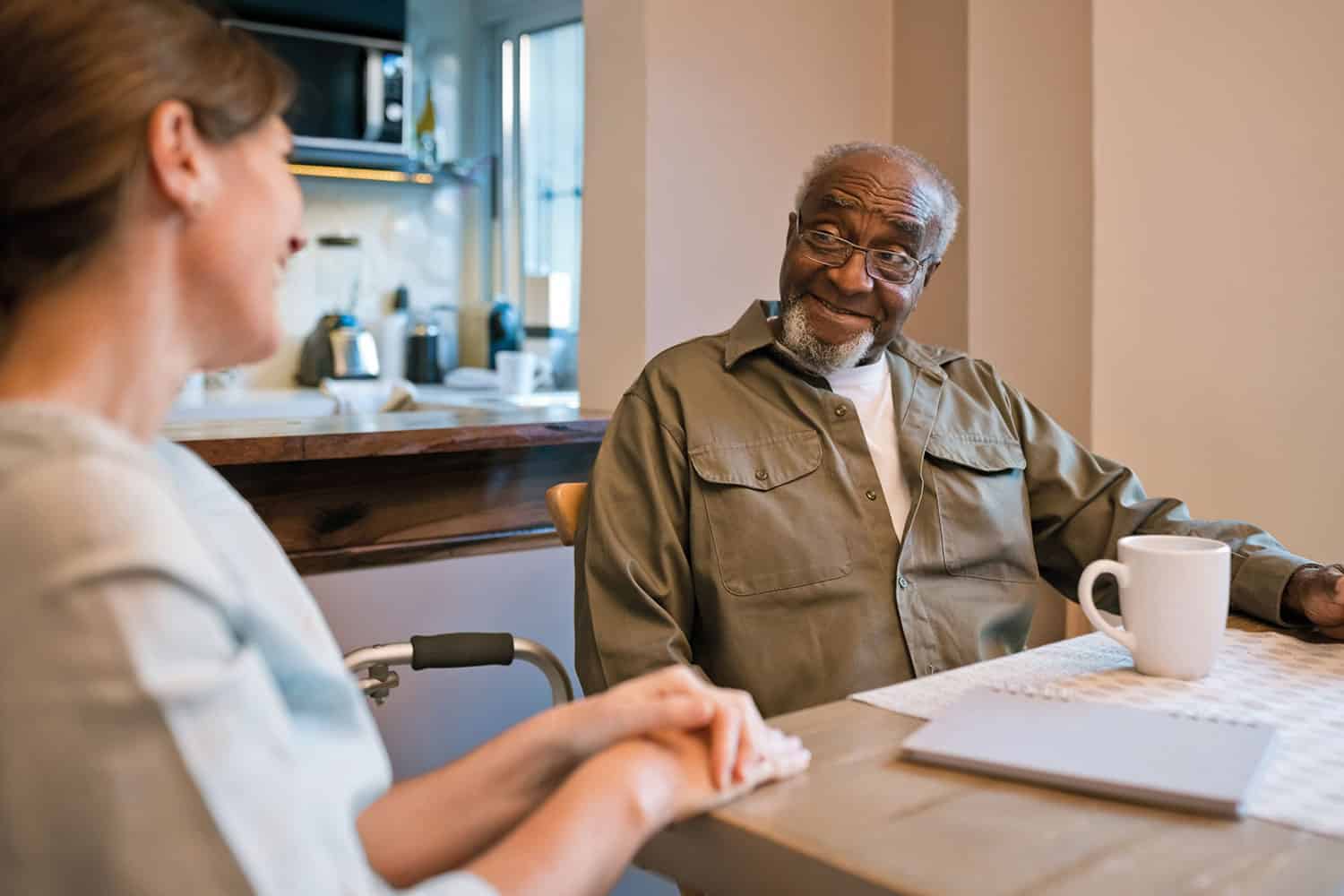 photo of side-view of woman carer having a coffee at a kitchen table with an older man