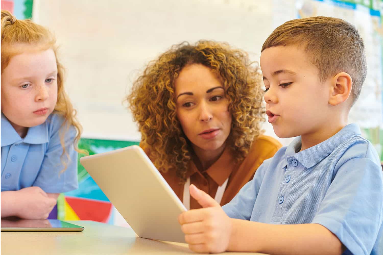 young boy reading to teacher from a tablet, with another child watching from the other side of the table, with a colourful wall display behind them