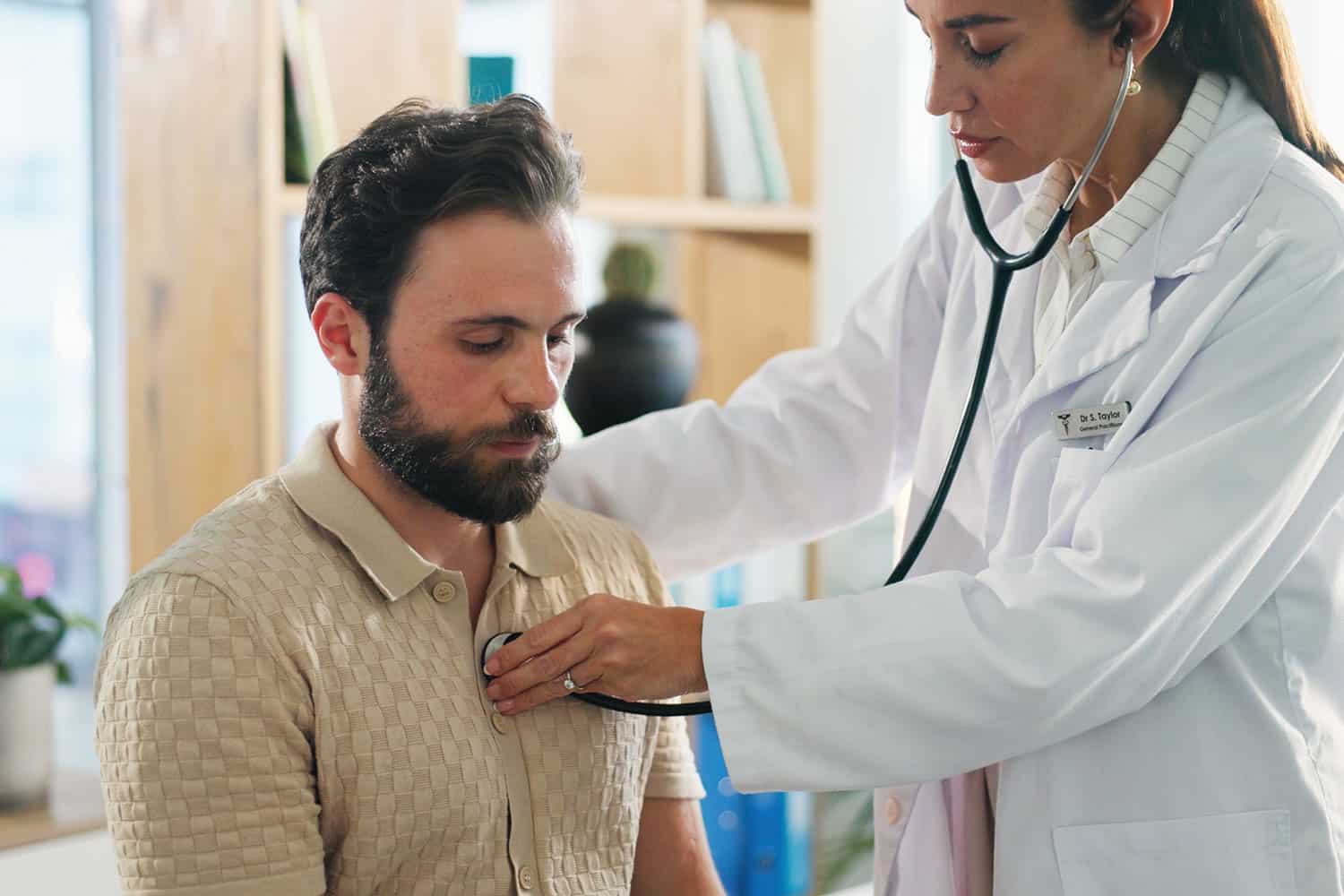 bearded man sitting down with a female doctor standing and holding a stethoscope to his chest