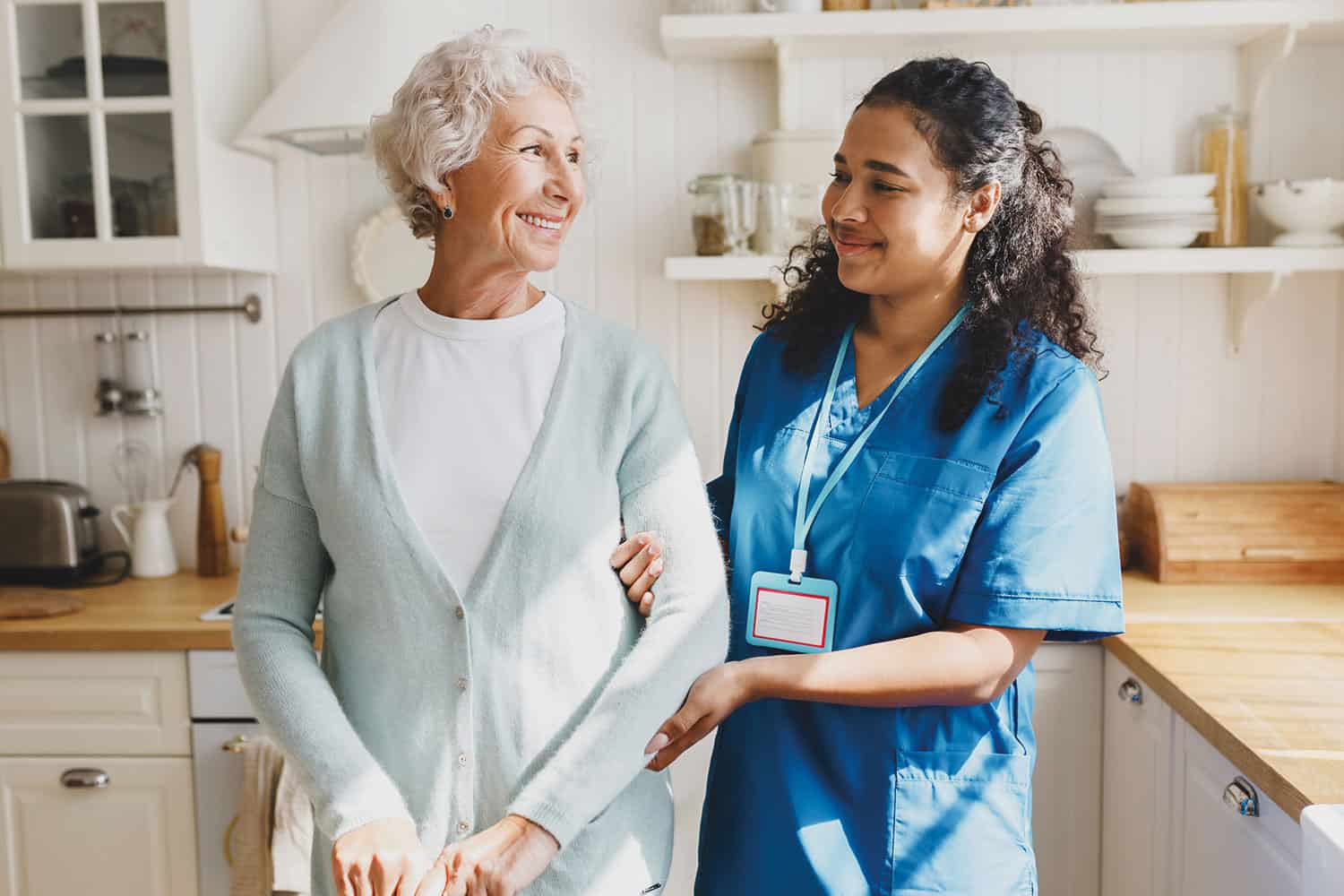 smiling older woman in her kitchen, arm-in-arm with uniformed carer