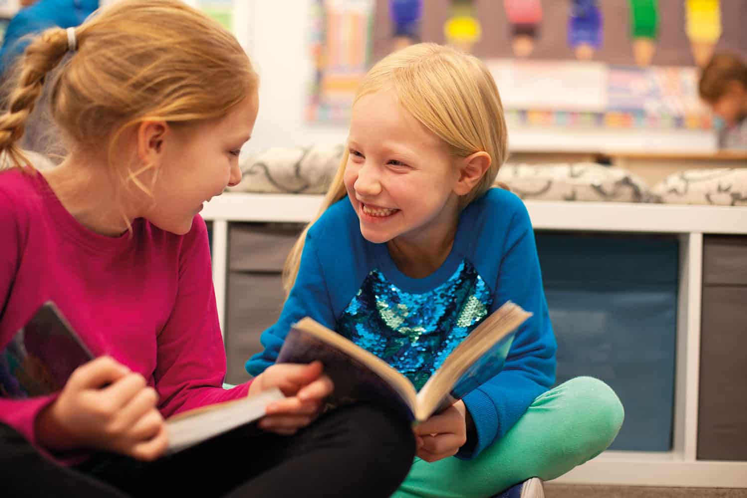 photo of two young girls sitting on floor reading and grinning at each other