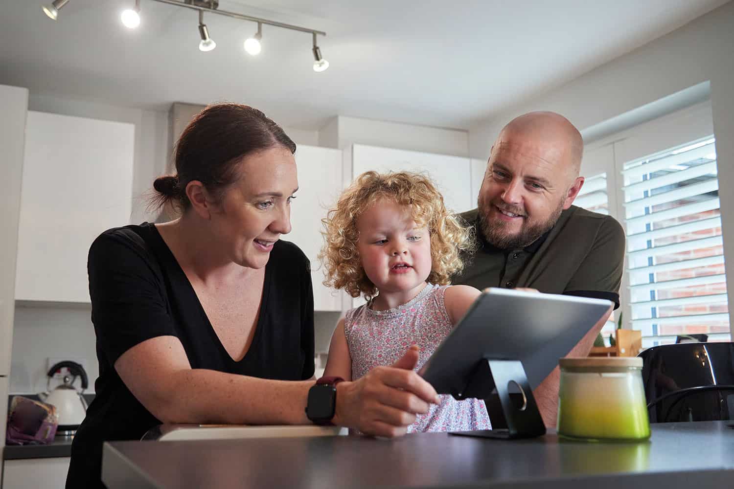 mum and dad at kitchen table with toddler daughter pointing at tablet