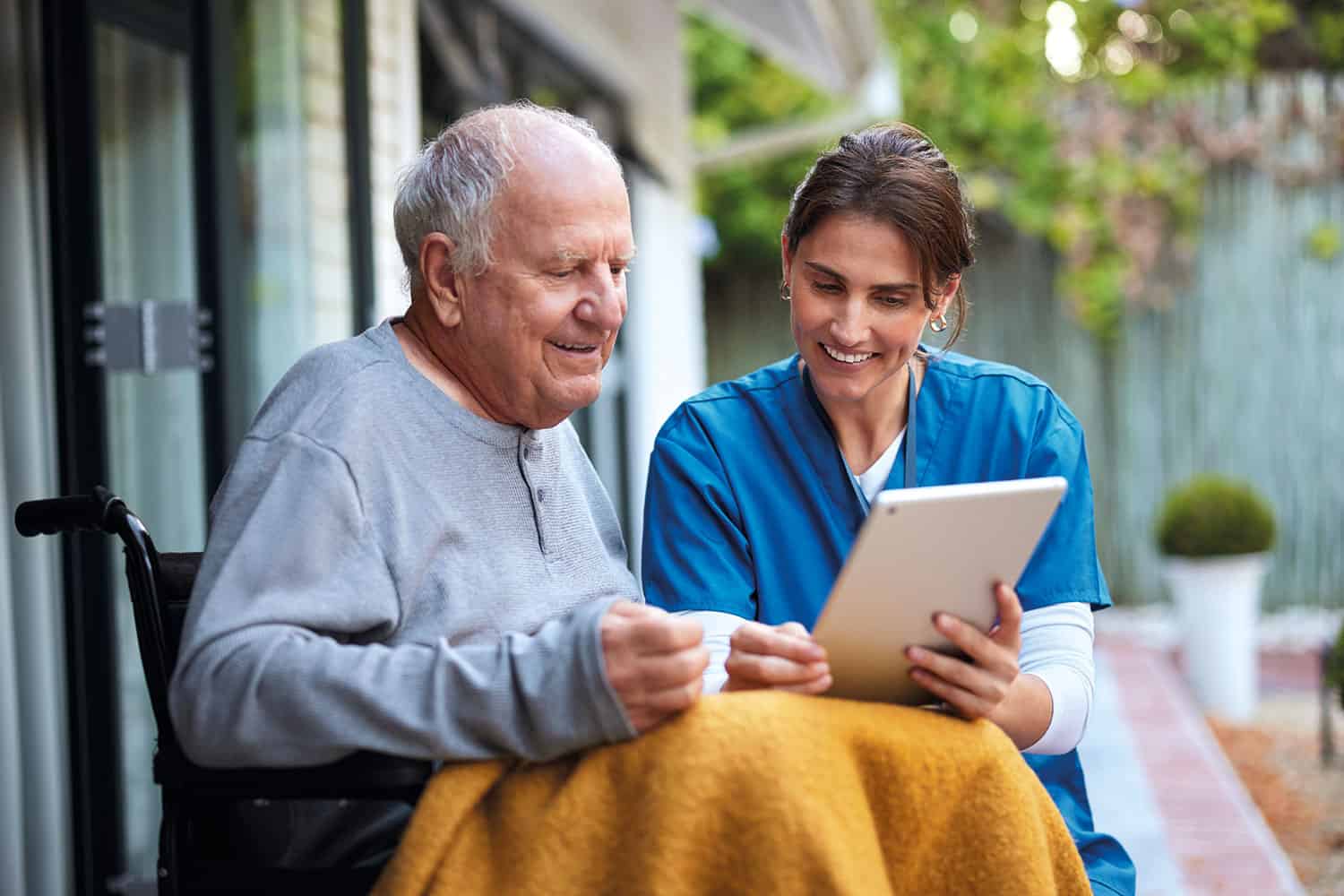 photo of older man sat in wheelchair smiling and looking at tablet with younger female carer