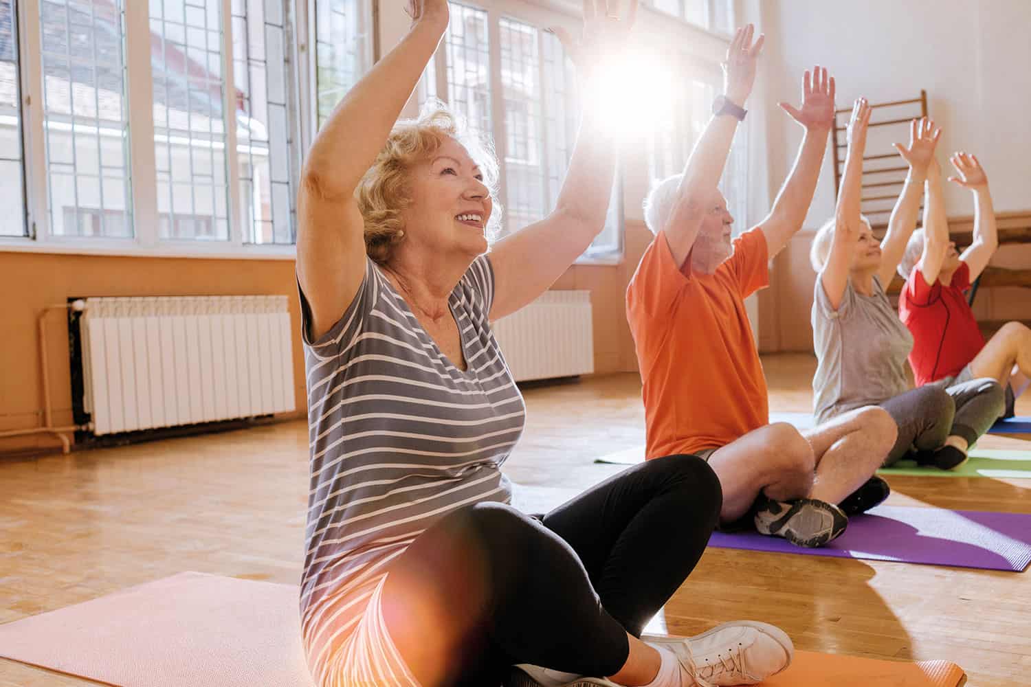 row of older men and women sitting on exercise mats with arms stretched up in sunny gym